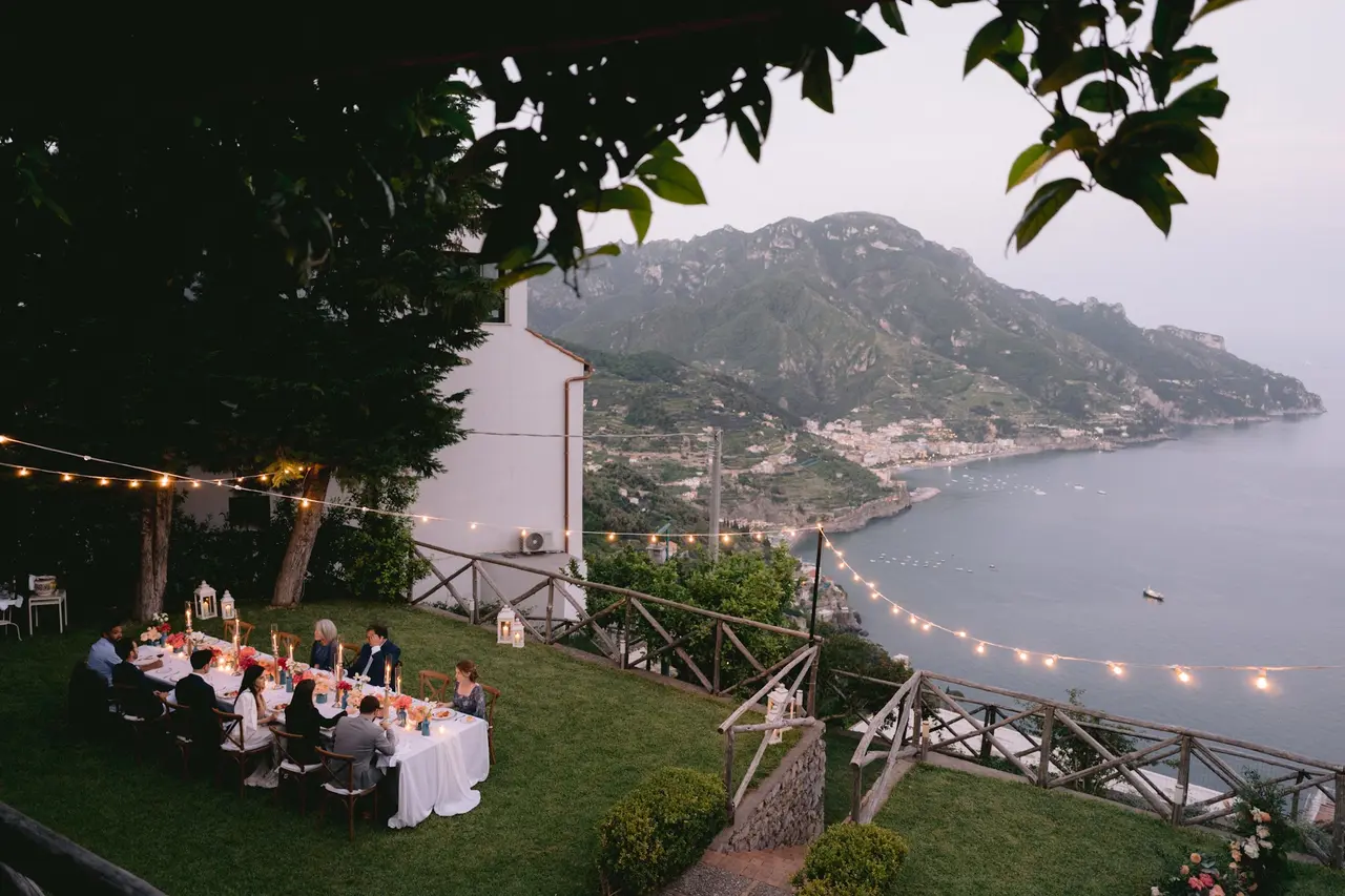 Guests seated at the long wedding table at Villa Mariella after sunset, lanterns and festoon lights above the coast